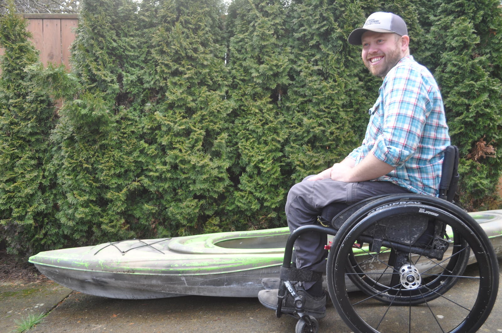White man wearing a baseball cap and a blue flannel shirt sitting in a wheelchair beside a green and black kayak. Behind him is a wall of green shrubbery.